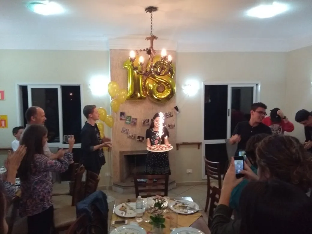Festa de aniversário com balões dourados, bolo e convidados reunidos em volta da mesa, em ambiente interno.