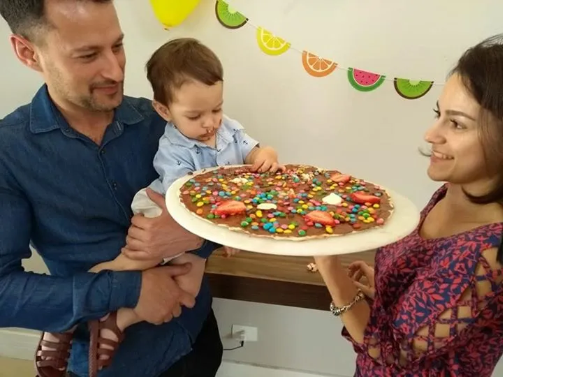 Família sorrindo em uma sala decorada, segurando uma pizza grande em uma bandeja.