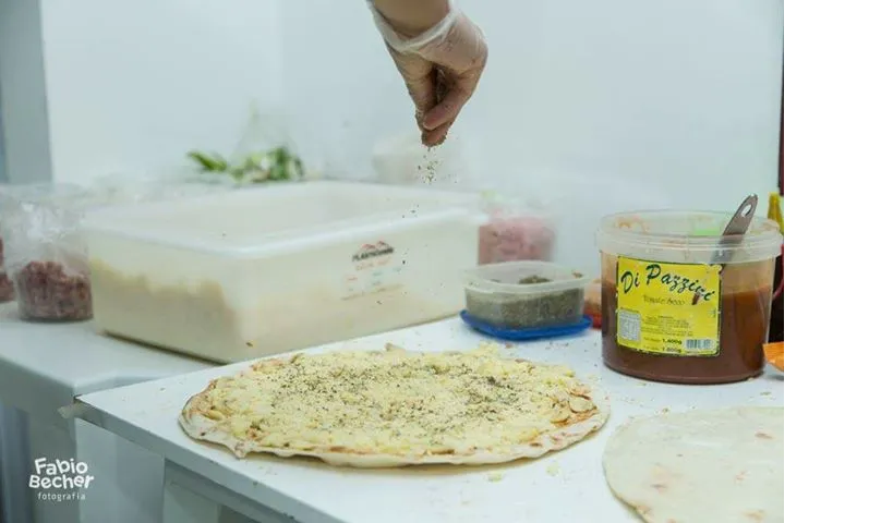 Tempero aplicado manualmente em um pão achatado ao lado de recipientes com ingredientes em uma bancada de cozinha.