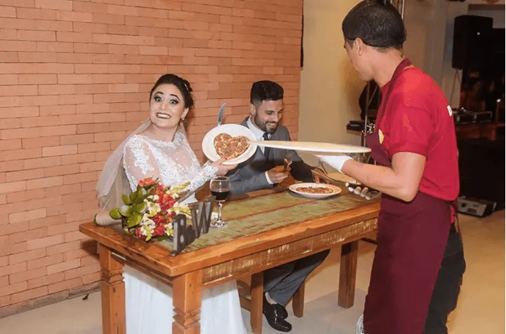 Noiva sentada à mesa decorada, sorrindo enquanto um garçom apresenta a comida em um salão de banquetes.