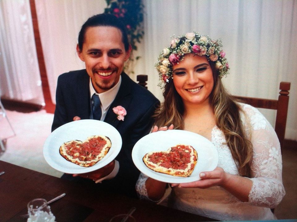 Casal sorridente à mesa segurando pratos de comida em formato de coração em um cenário de casamento.