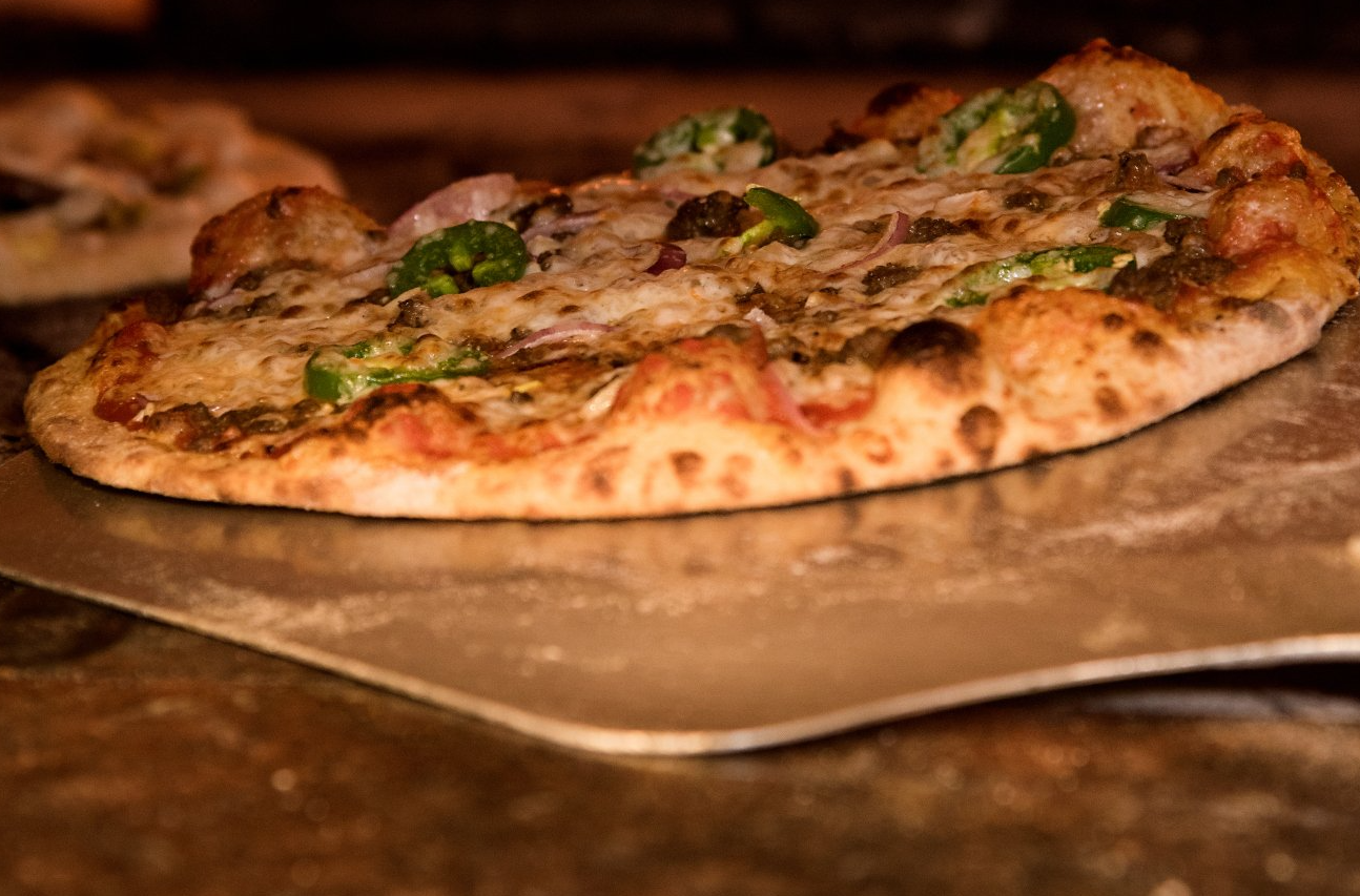 Pizza recém-assada com cobertura verde em uma pá de metal em uma área de forno com pouca luz.