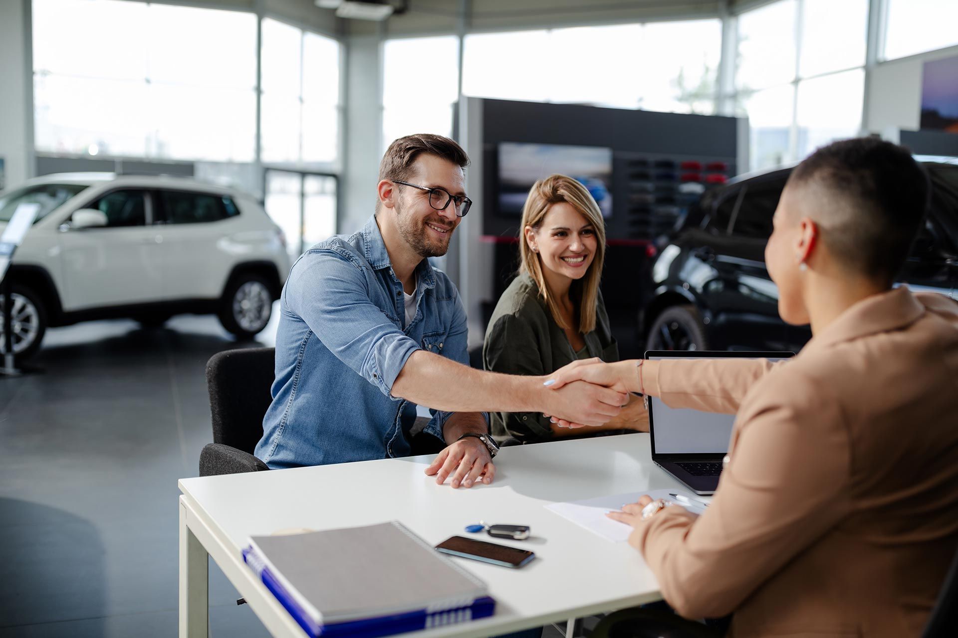 Car salesperson shakes hands with a customer in a bright showroom, with a woman smiling nearby. Car salesperson shakes hands with a customer in a bright showroom, with a woman smiling nearby.