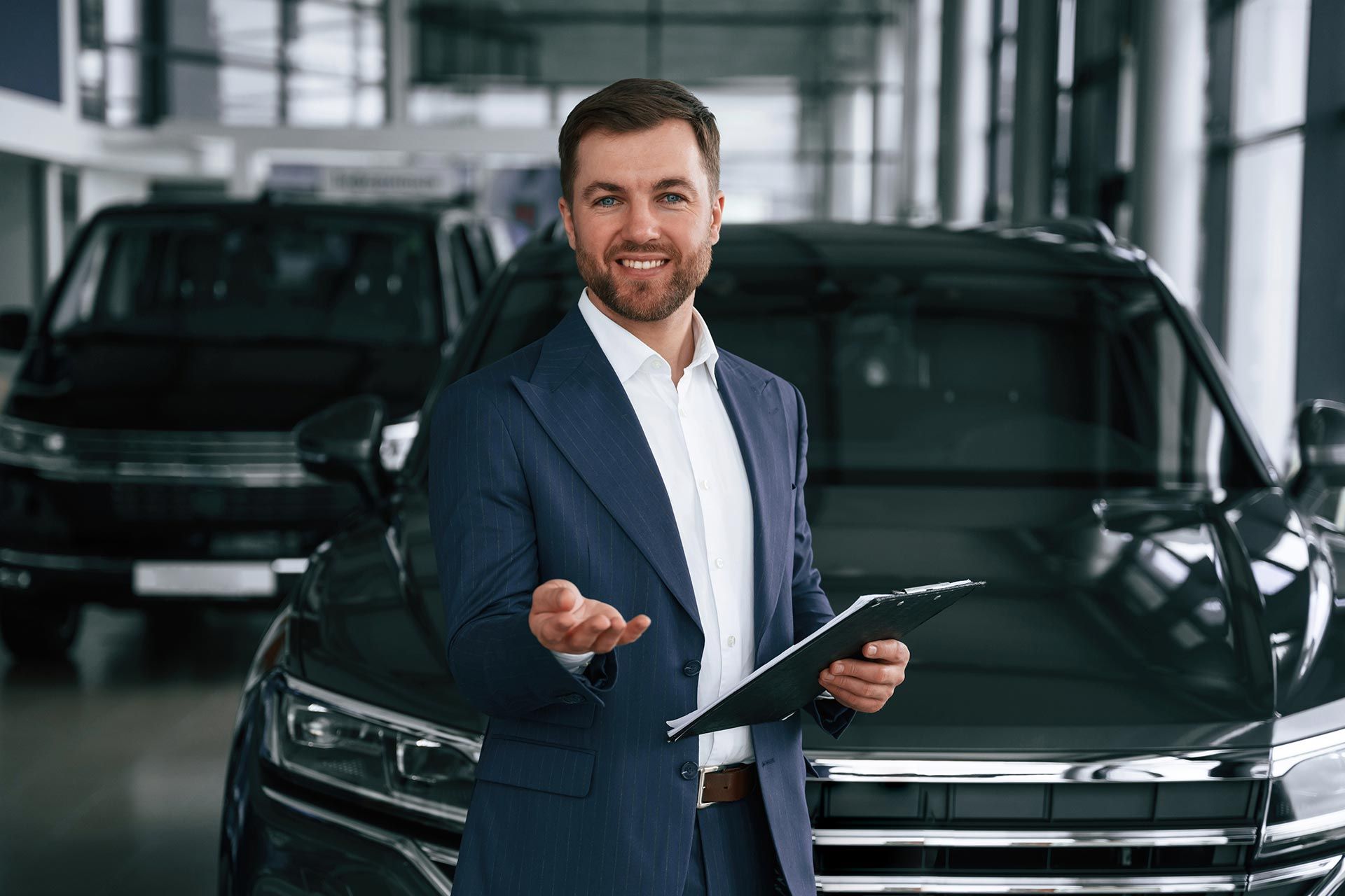 Salesman in a navy suit holding a clipboard in a car showroom, standing beside a black SUV