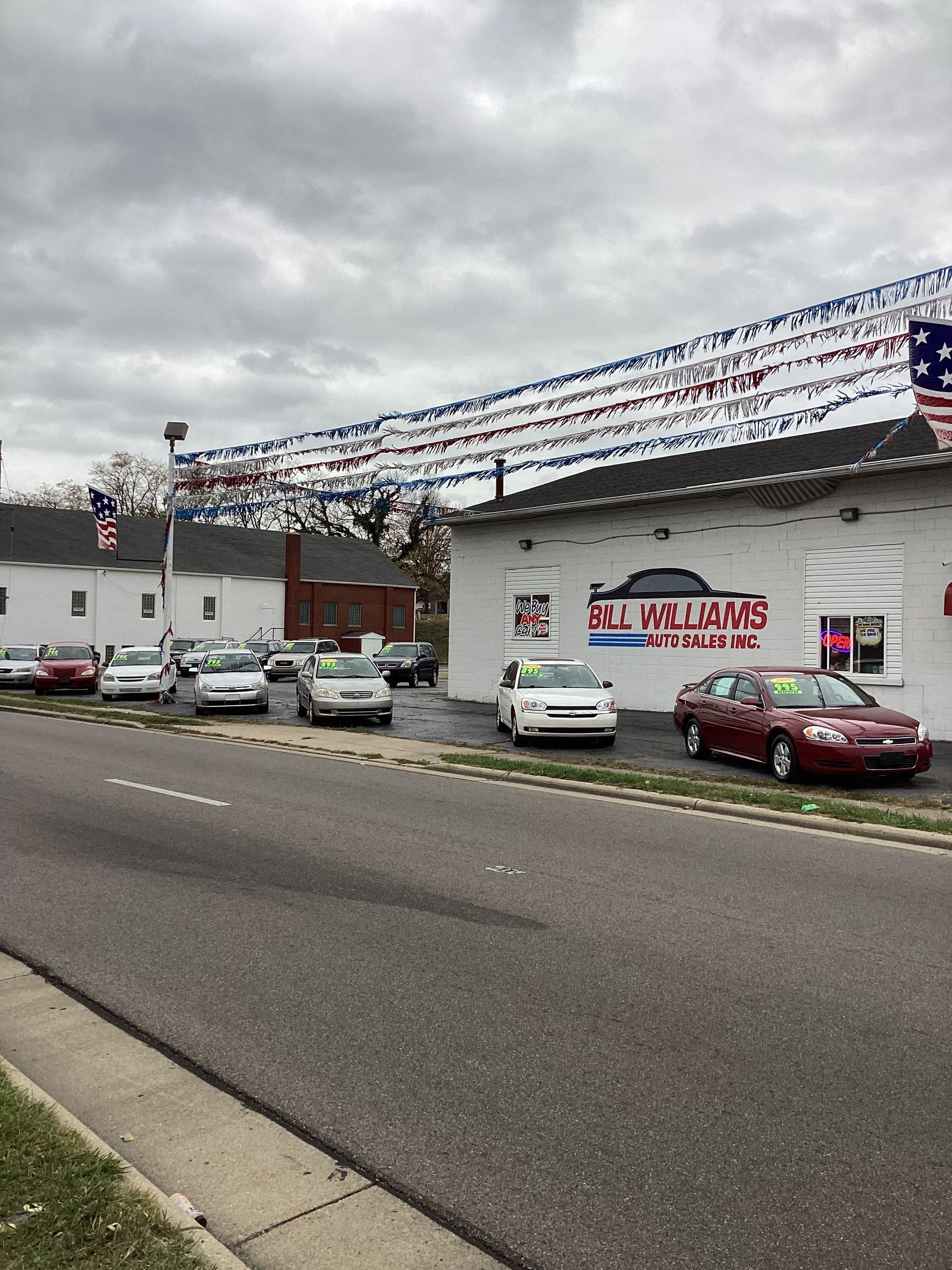 Car dealership lot with several cars parked in front of a white building under a cloudy sky.