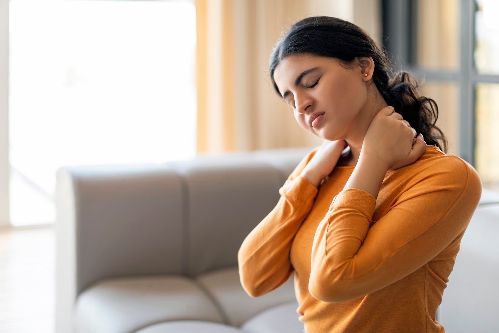Woman in an orange sweater massaging her neck on a couch, looking down in discomfort