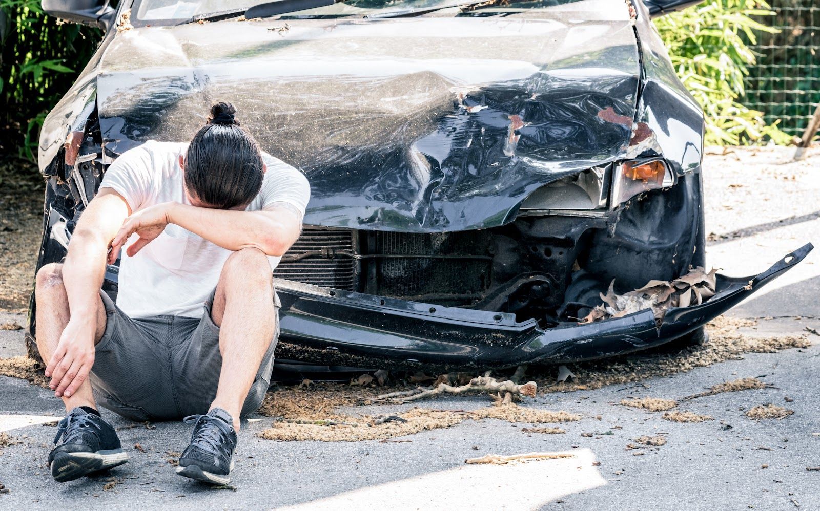 Man sitting on the ground beside a heavily damaged black car after a crash, head in hand.