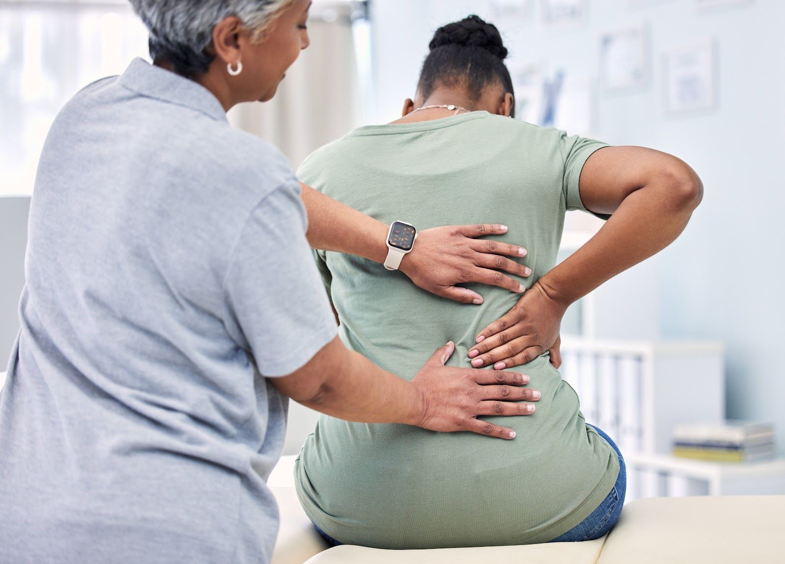 Therapist supports a seated patient holding her lower back in a bright clinic room