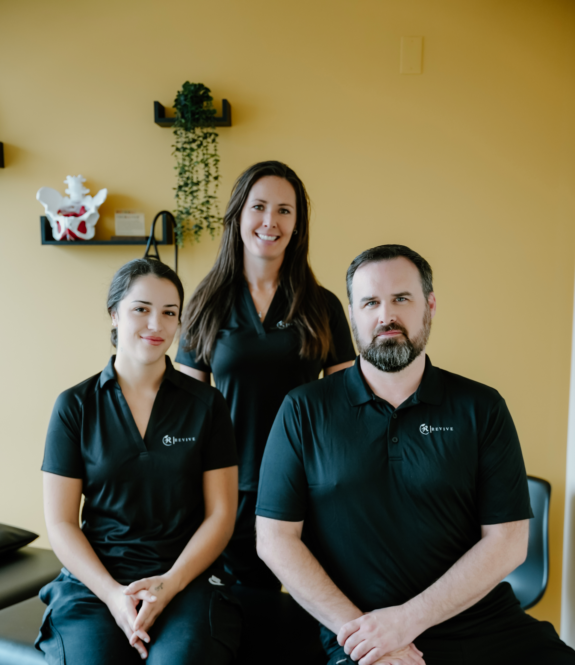 Three people in black polos pose indoors against a yellow wall, smiling at the camera.