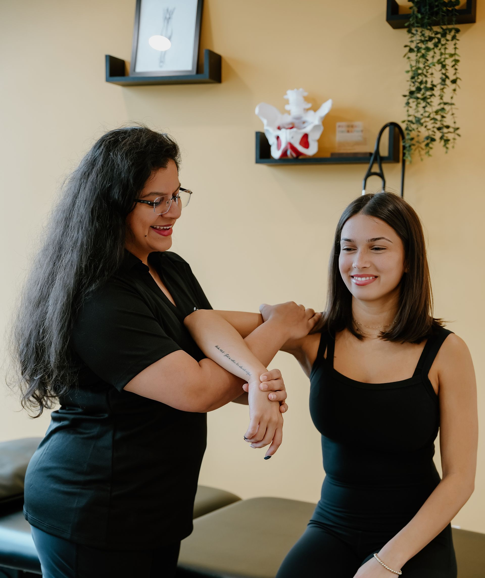 Two women smiling during an arm massage in a cozy room with shelves and plants.