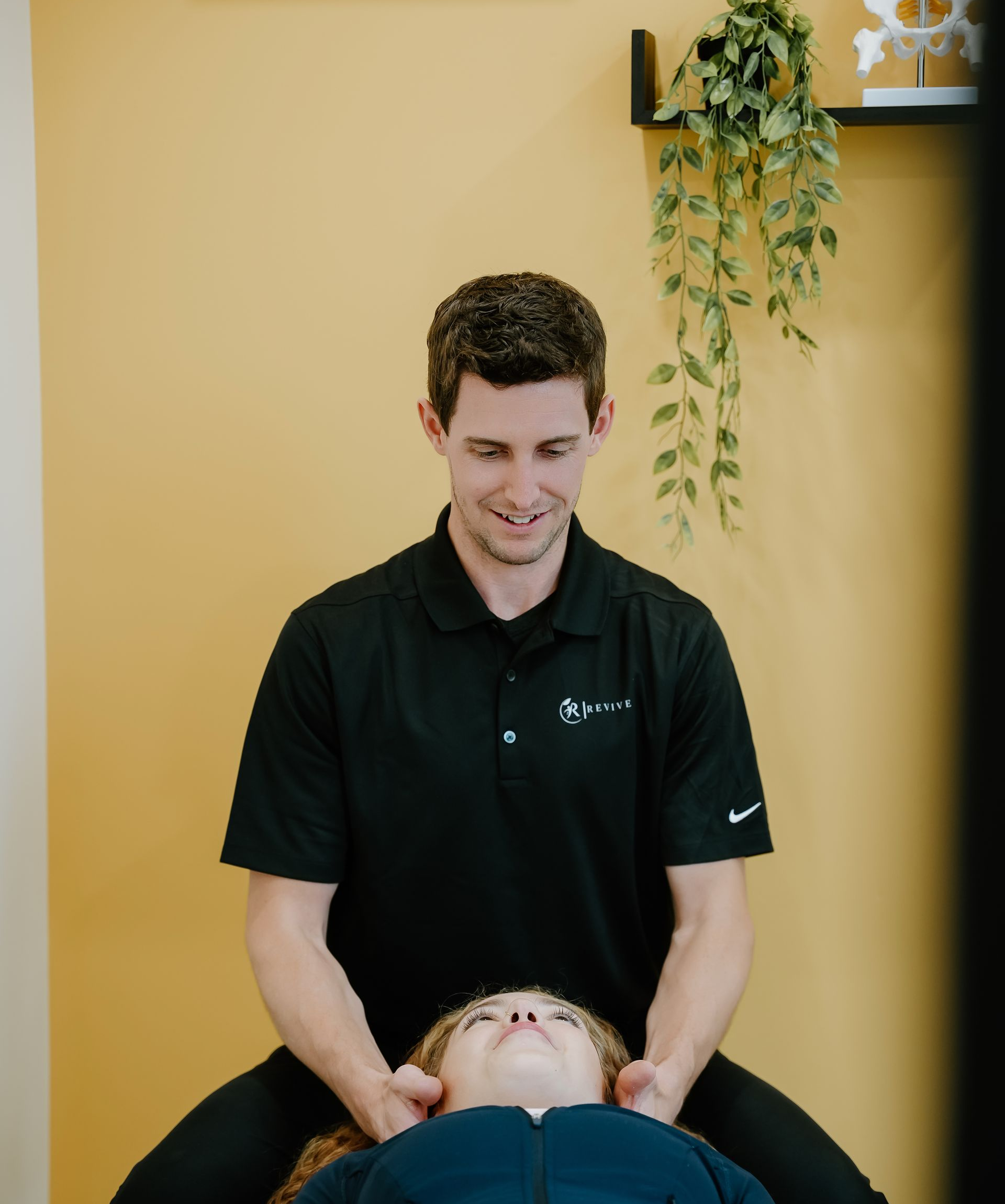 Chiropractor in black polo gently adjusts a patient’s neck in a clinic with a yellow wall.