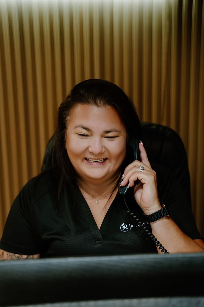 Woman in a black shirt smiling while talking on a phone at a desk, with vertical wood panels behind her.