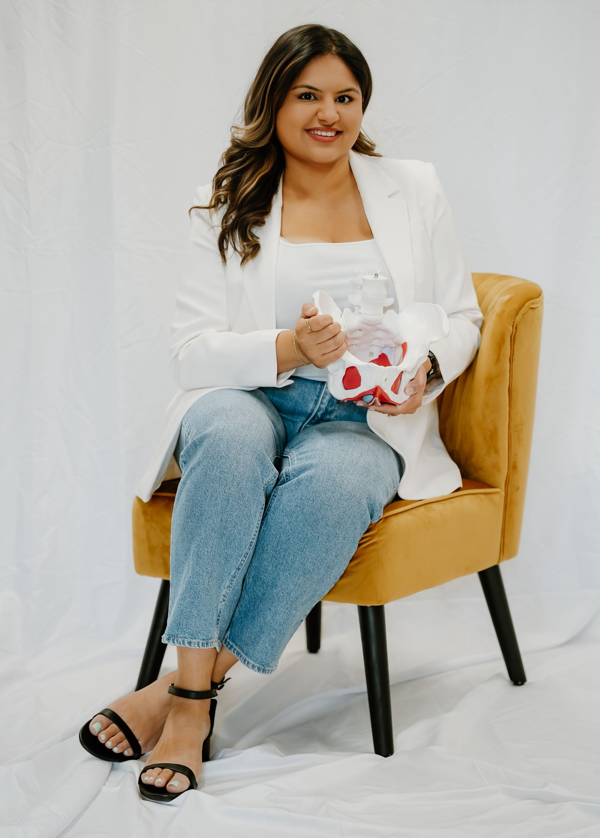 Woman seated on mustard chair, wearing white blazer and jeans, holding a white mug with red hearts.