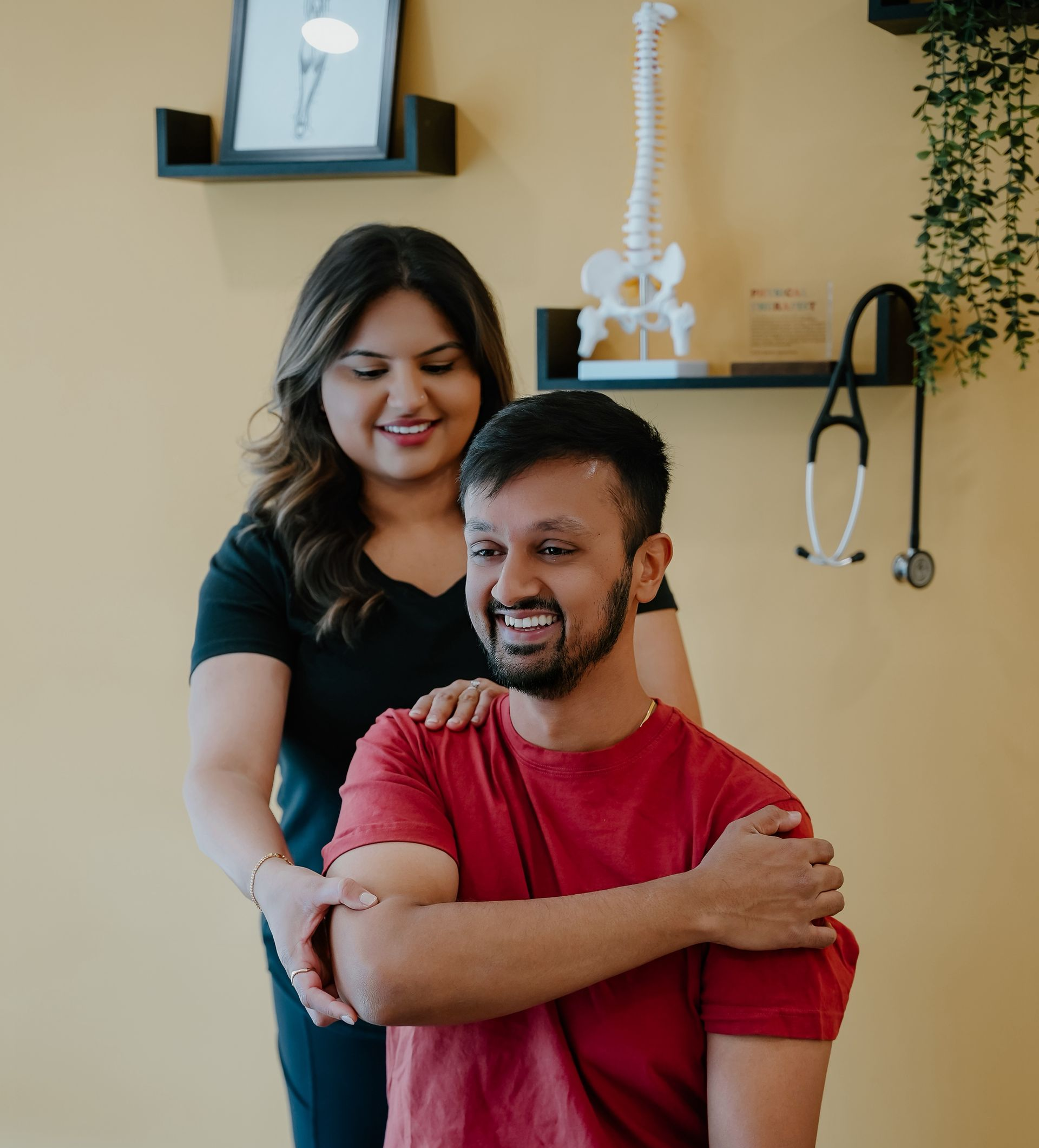 Two people smiling in a clinic, one seated in a red shirt while the other stands behind with a hand on his shoulder.