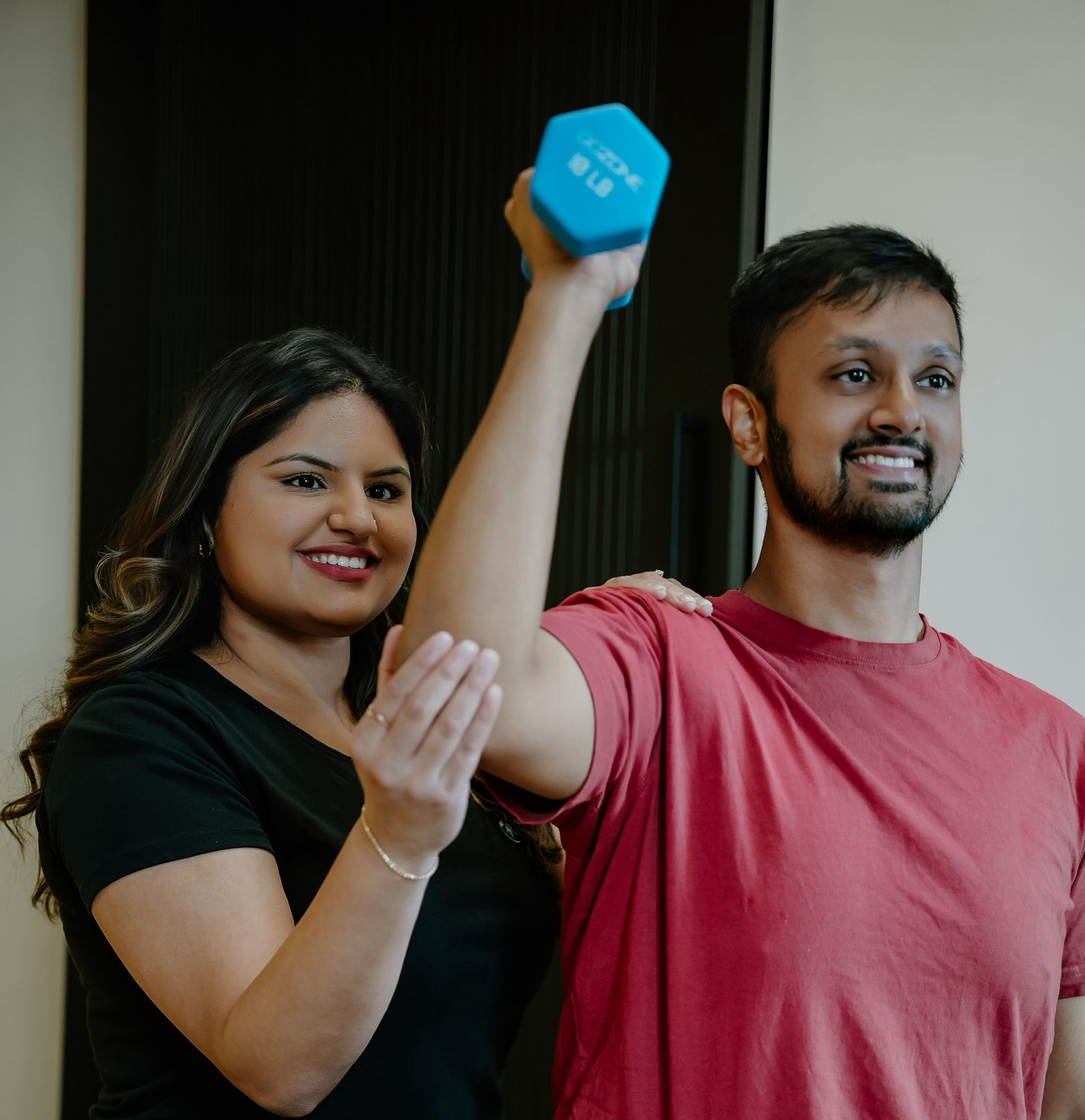 Two people posing indoors, one holding a blue dumbbell overhead while the other claps nearby.