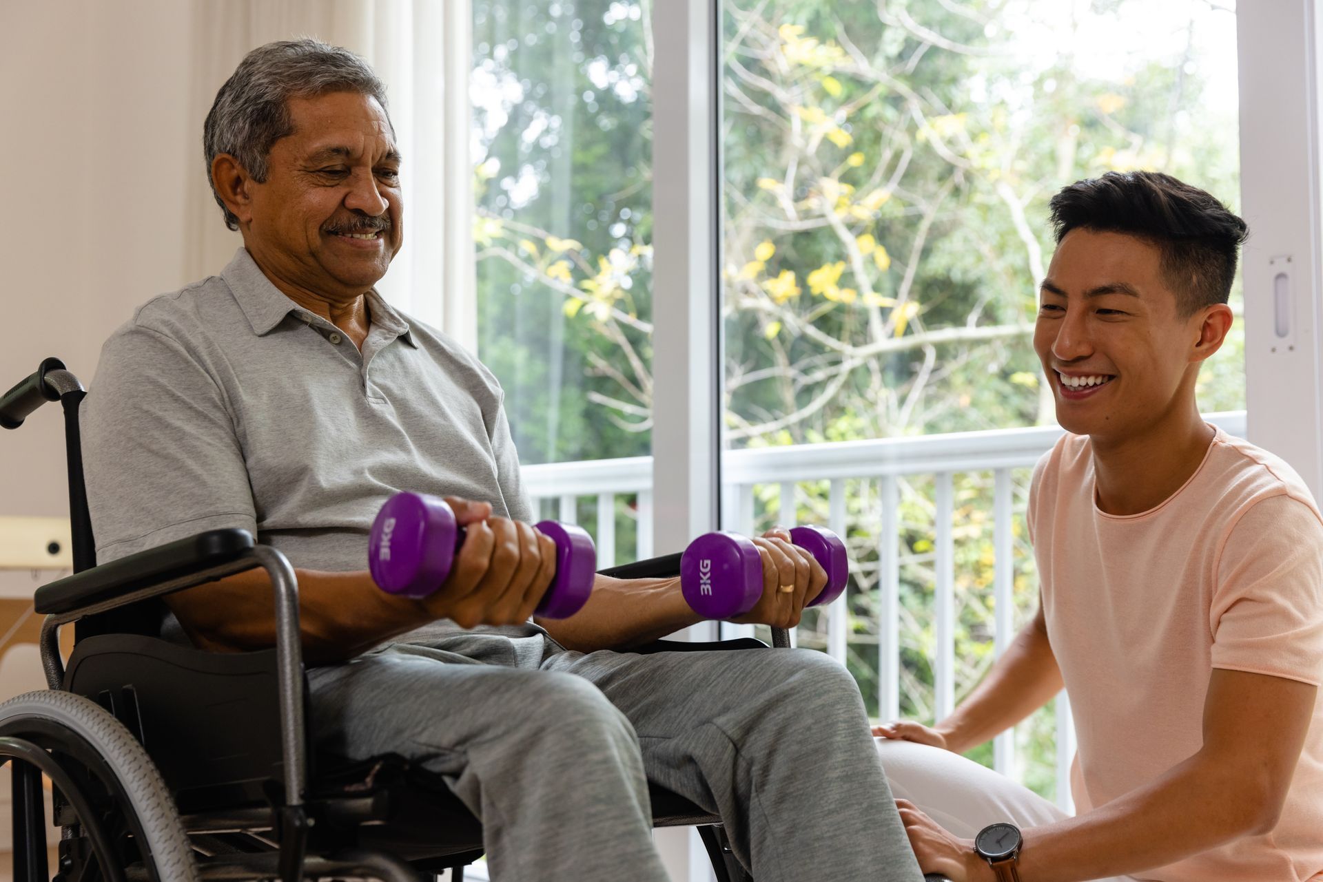 Two people smiling during seated arm exercise with purple dumbbells in a bright room