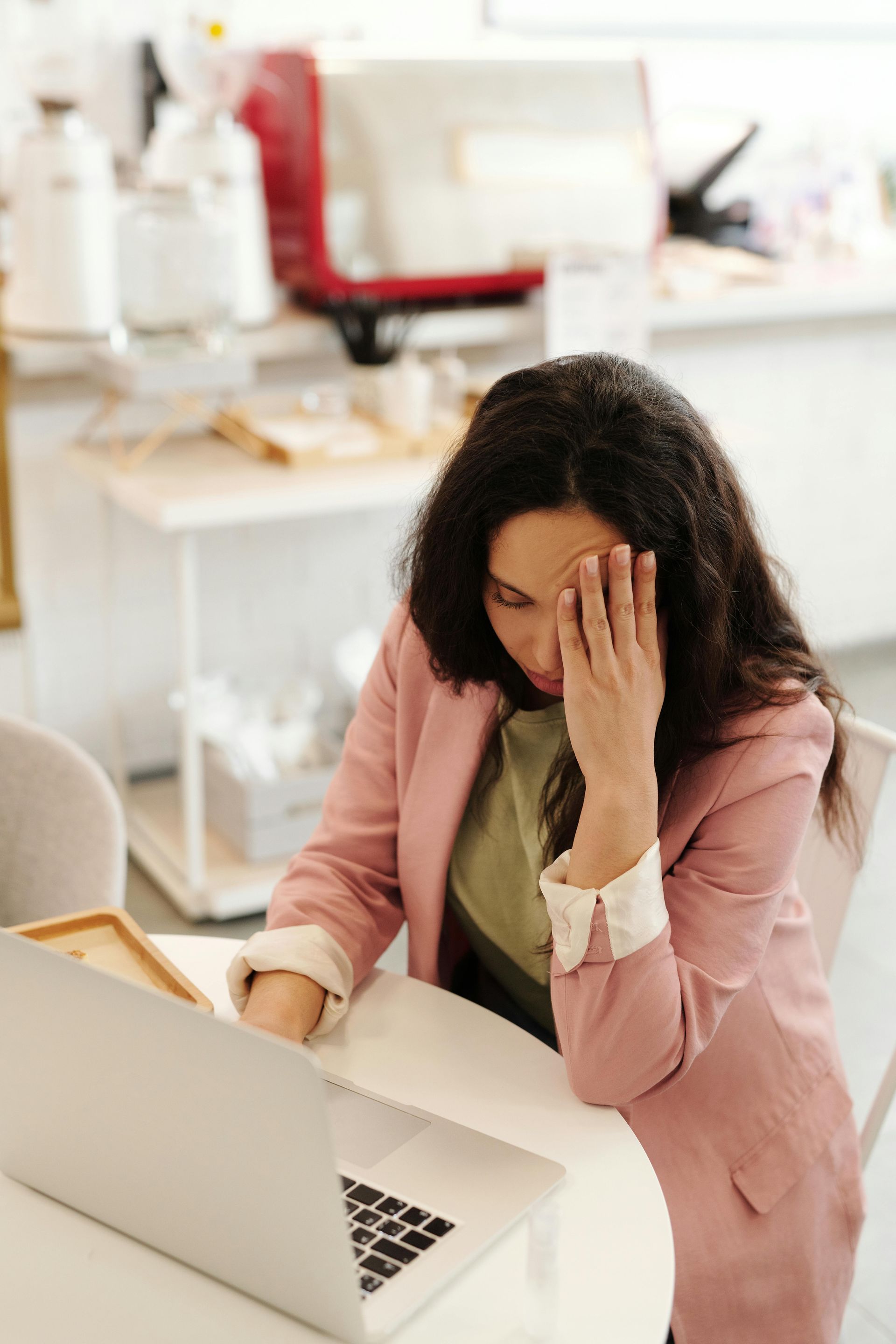 Woman in a pink blazer sits at a laptop, holding her forehead in a bright office.
