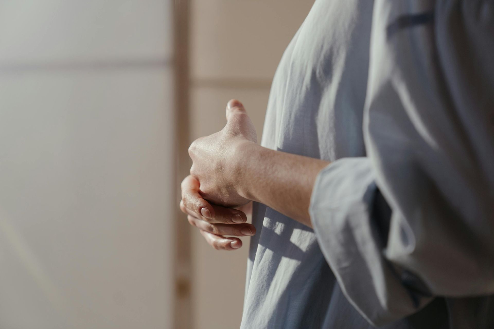 Hands clasped in front of a gray shirt, with a blurred indoor background.