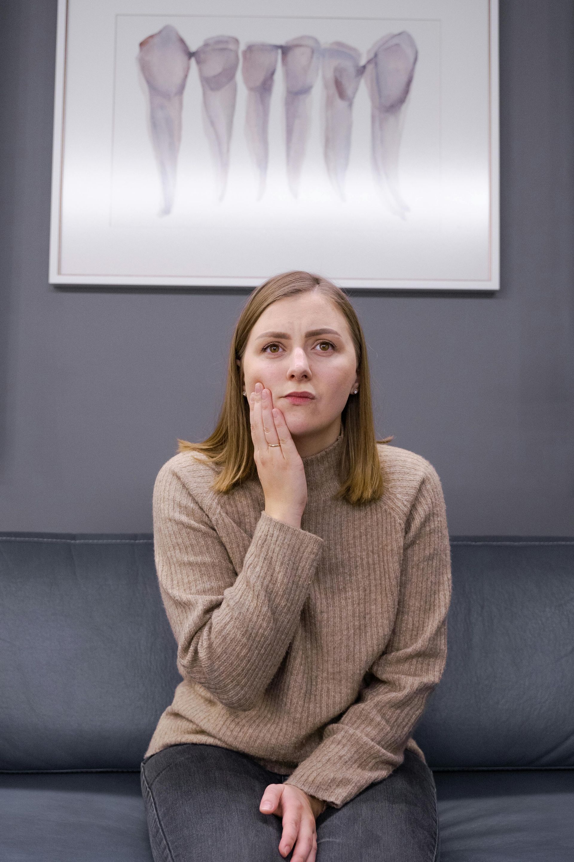 Woman sitting on a couch with hand on cheek, looking thoughtful, in front of a framed dental x-ray.