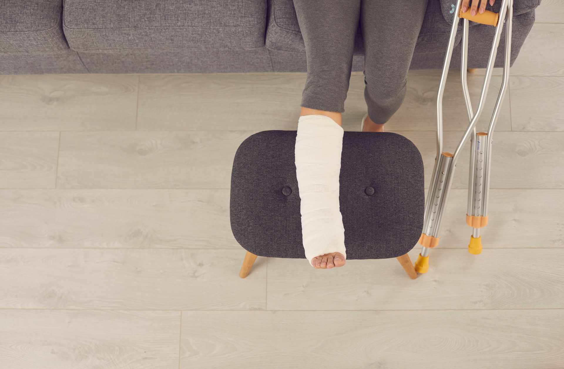 Person with a leg cast using crutches, standing beside a black ottoman on a light wood floor.