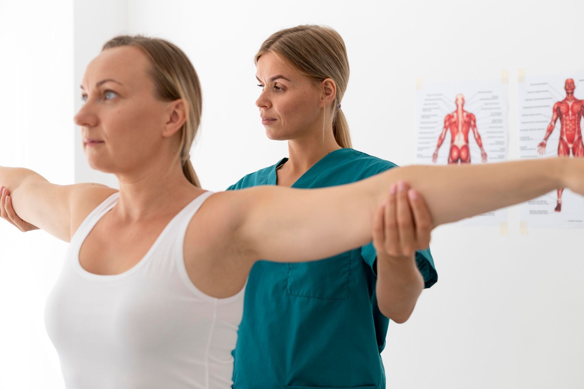 Physical therapist assisting patient with arm stretches in a clinic, with anatomy charts in the background