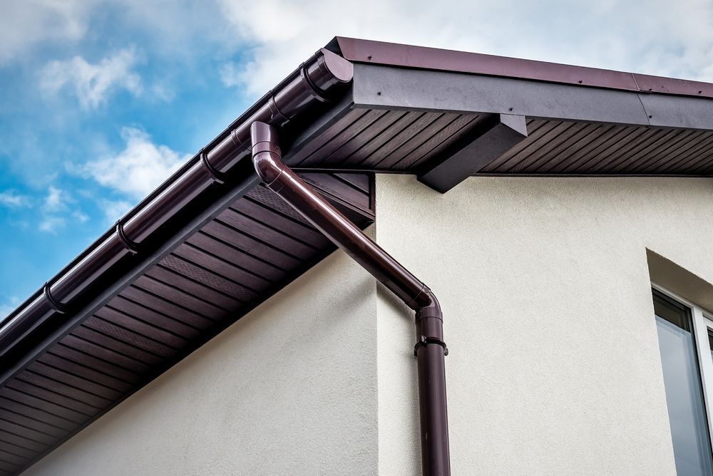 Brown gutters and siding on a white house against a blue sky with clouds.