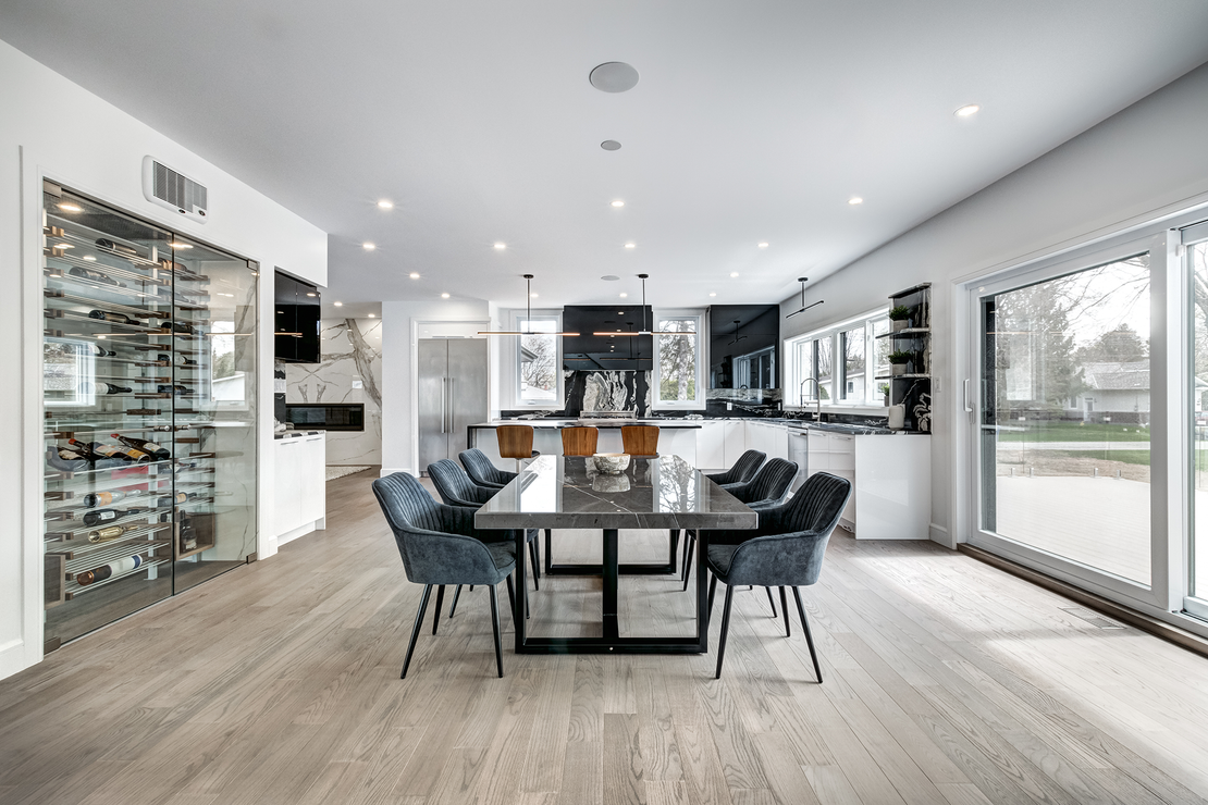 Modern dining room with grey table and blue chairs; kitchen in background; light wood floors.