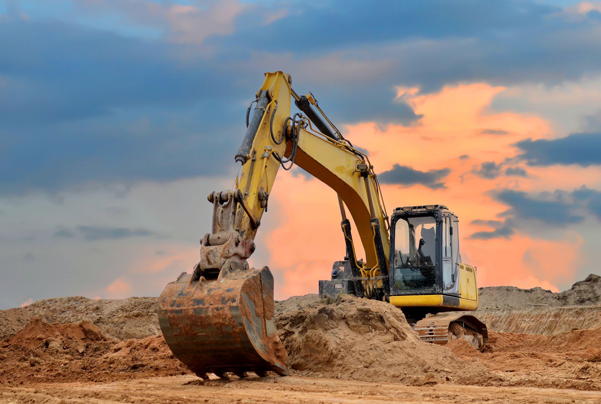 Yellow excavator digging soil at a construction site with a cloudy sunset sky in the background.