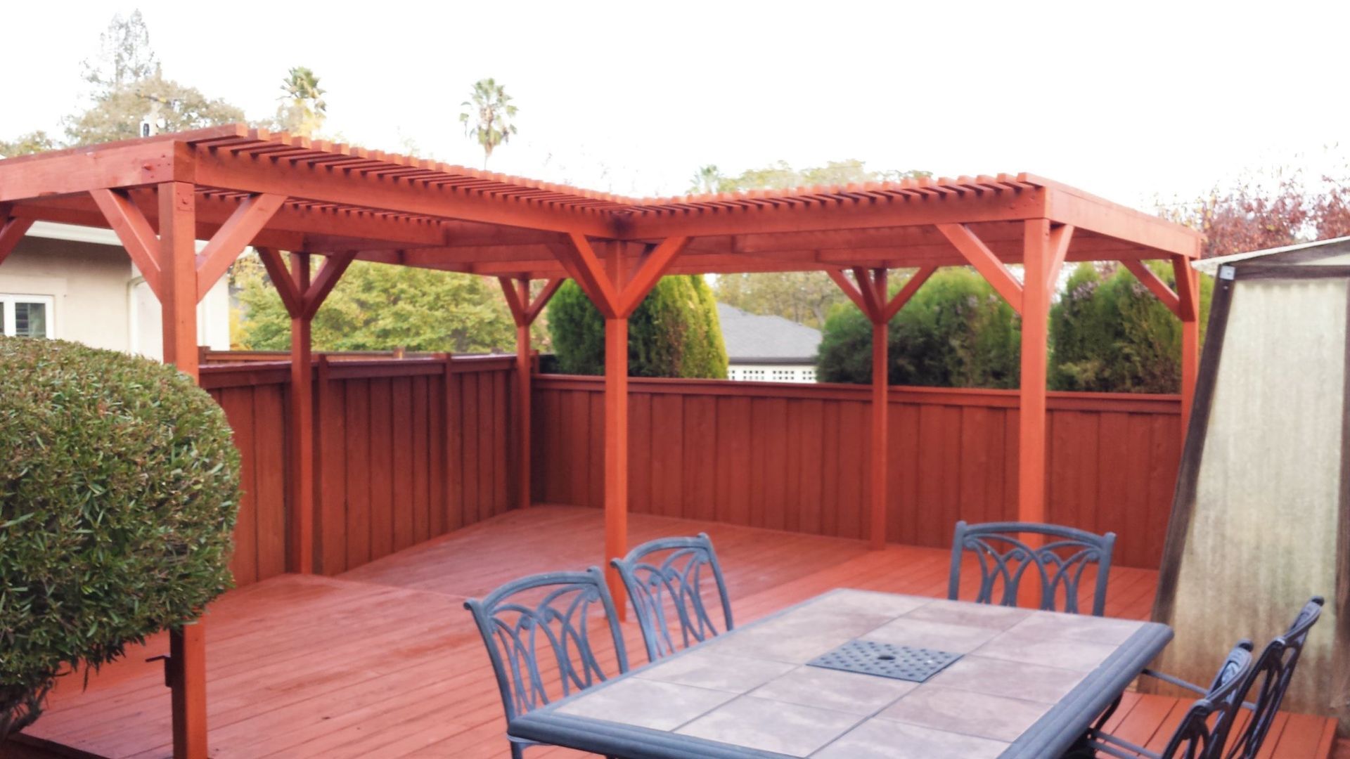 Red wooden pergola over a deck with a table and chairs, enclosed by a red fence.