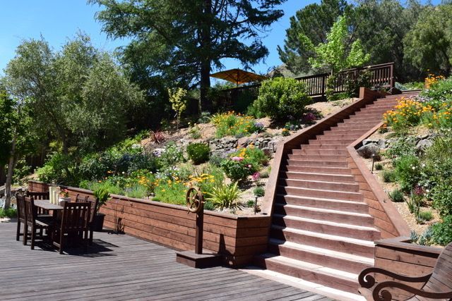 Wooden steps lead up a hillside garden with colorful flowers and trees. A patio area has a table and chairs.