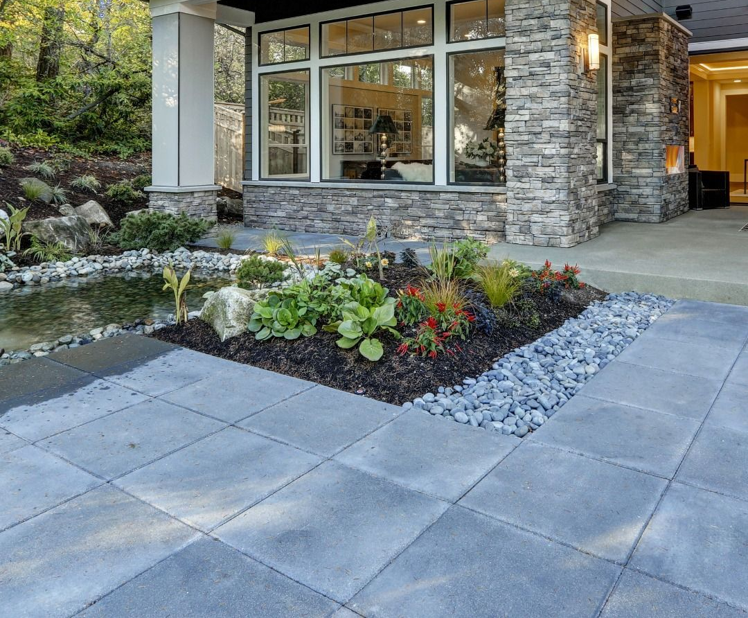 Gray paver patio with a garden bed and a stone-clad building facade. Pond to the left.