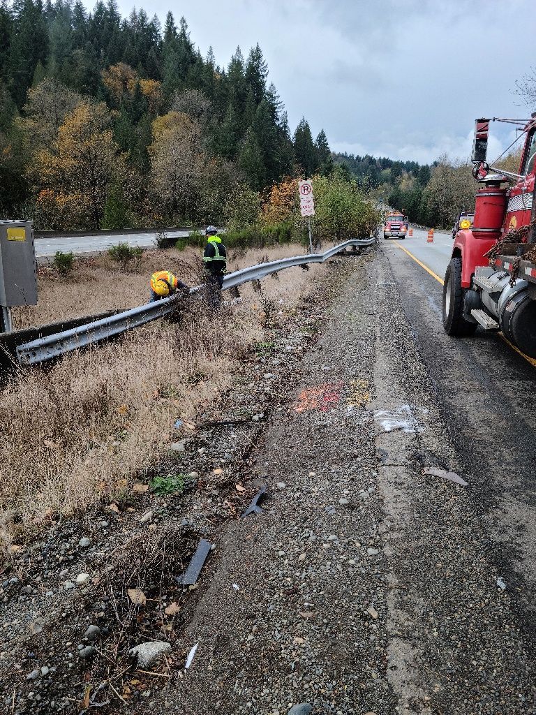Tow truck and workers near damaged guardrail on a roadside. Forest background, overcast sky.