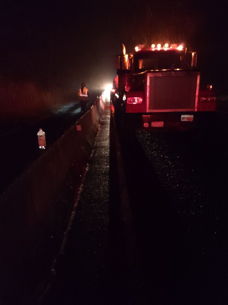 Semi-truck parked on a dark highway at night with emergency lights on. A person stands nearby.
