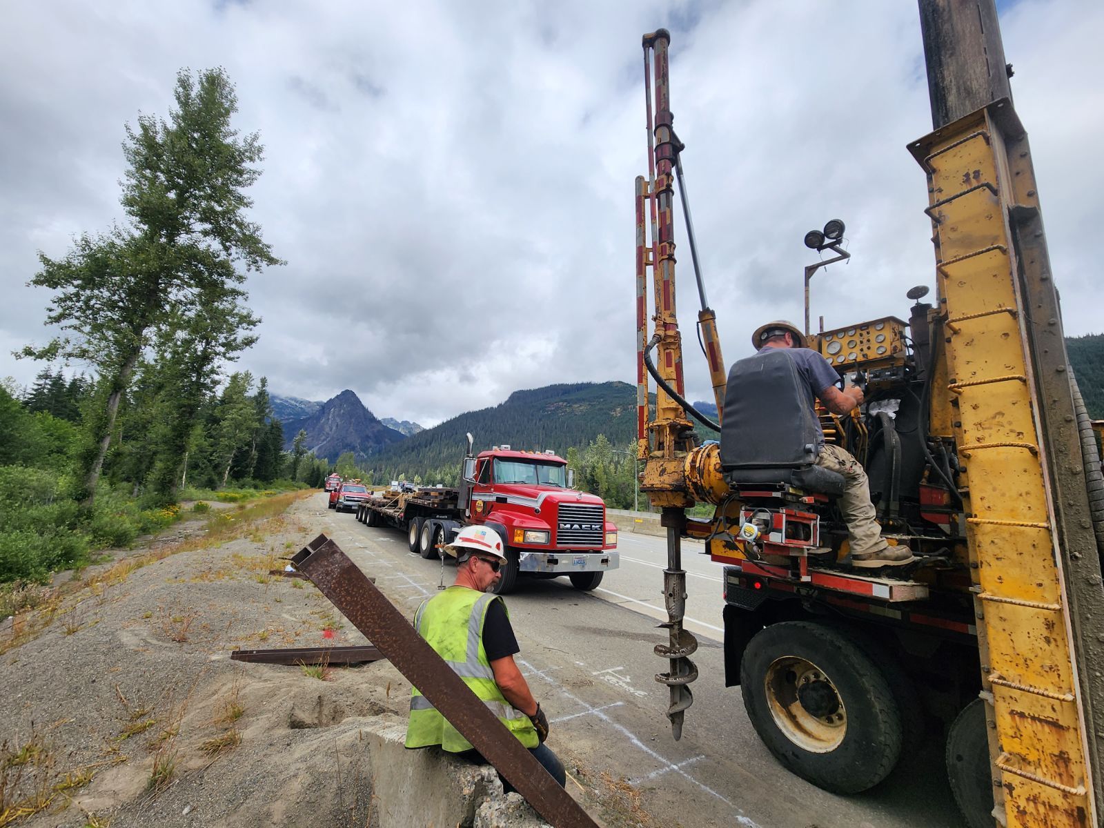 Construction workers operating a drilling machine on a road. Red truck, mountains, and trees in the background.