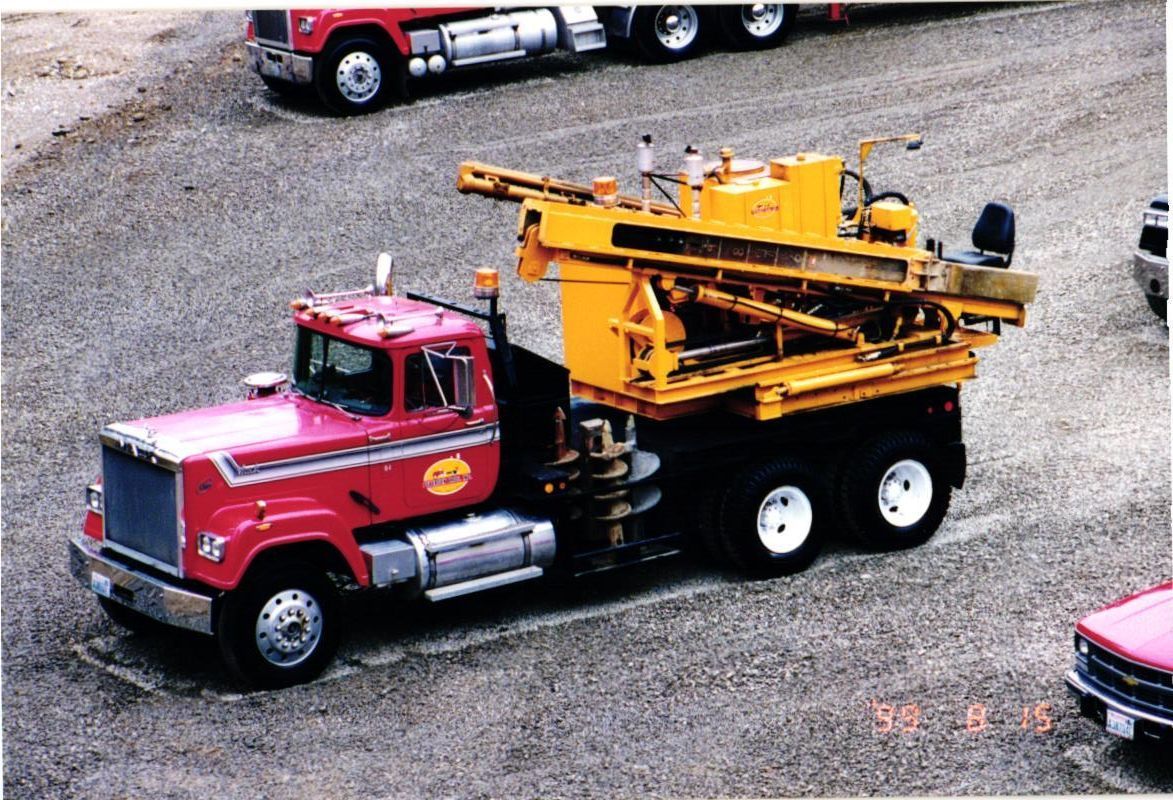 Red truck with a yellow drilling rig, parked on gravel.