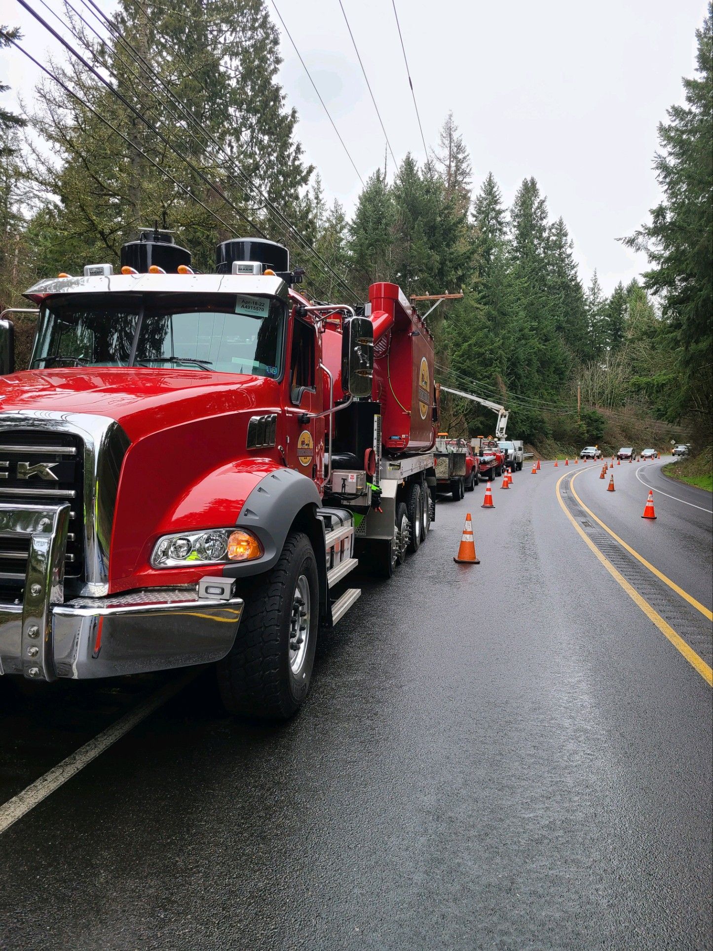 Red utility truck on a wet road, cones marking a work zone. Power lines and trees in the background.