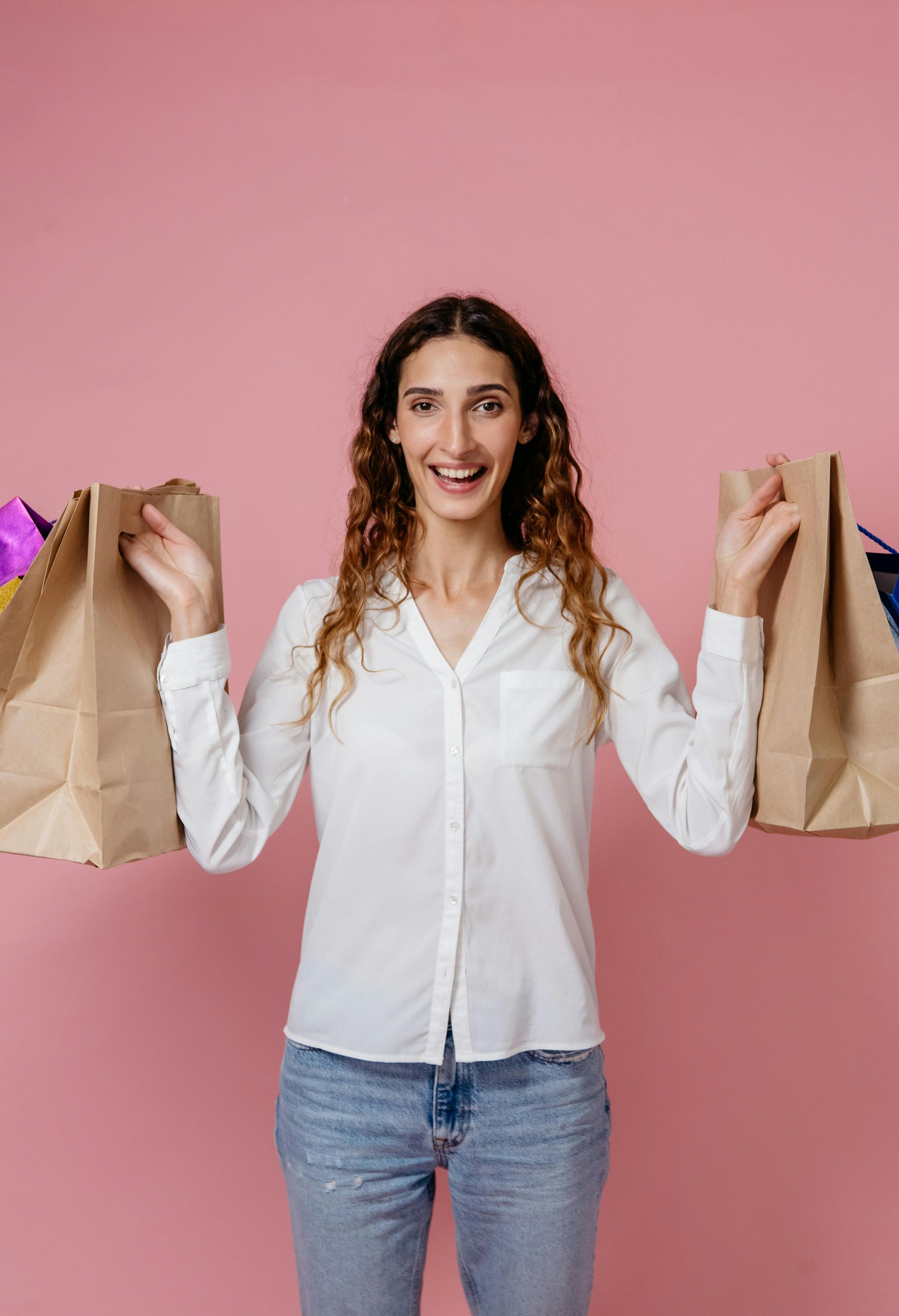 Uma mulher segura duas sacolas de compras nas mãos sobre um fundo rosa.
