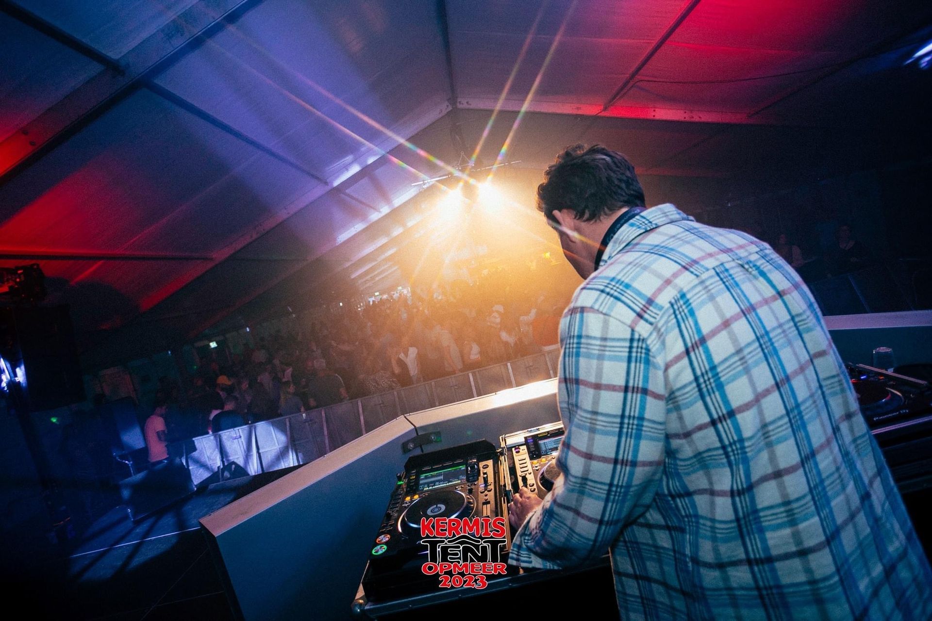 DJ draait achter een clubconsole onder fel podiumlicht en rood-blauwe plafondverlichting.