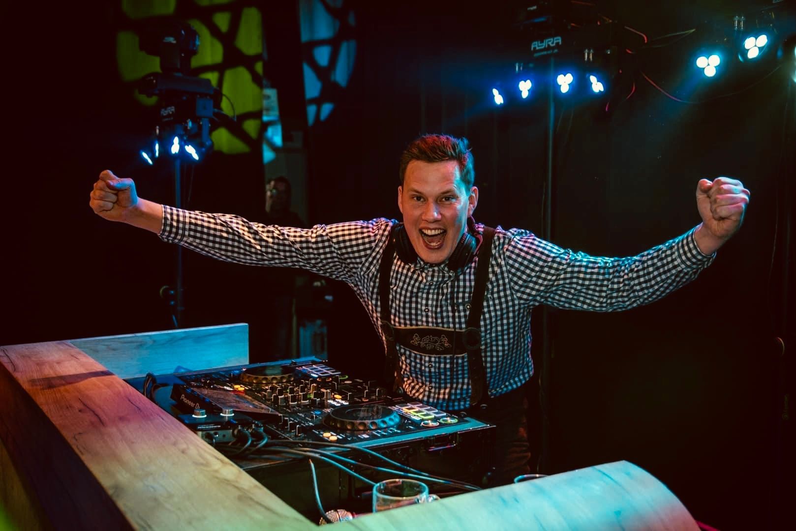 DJ in een geruit overhemd staat achter een mengpaneel te juichen op een podium dat verlicht is met neonlichten.