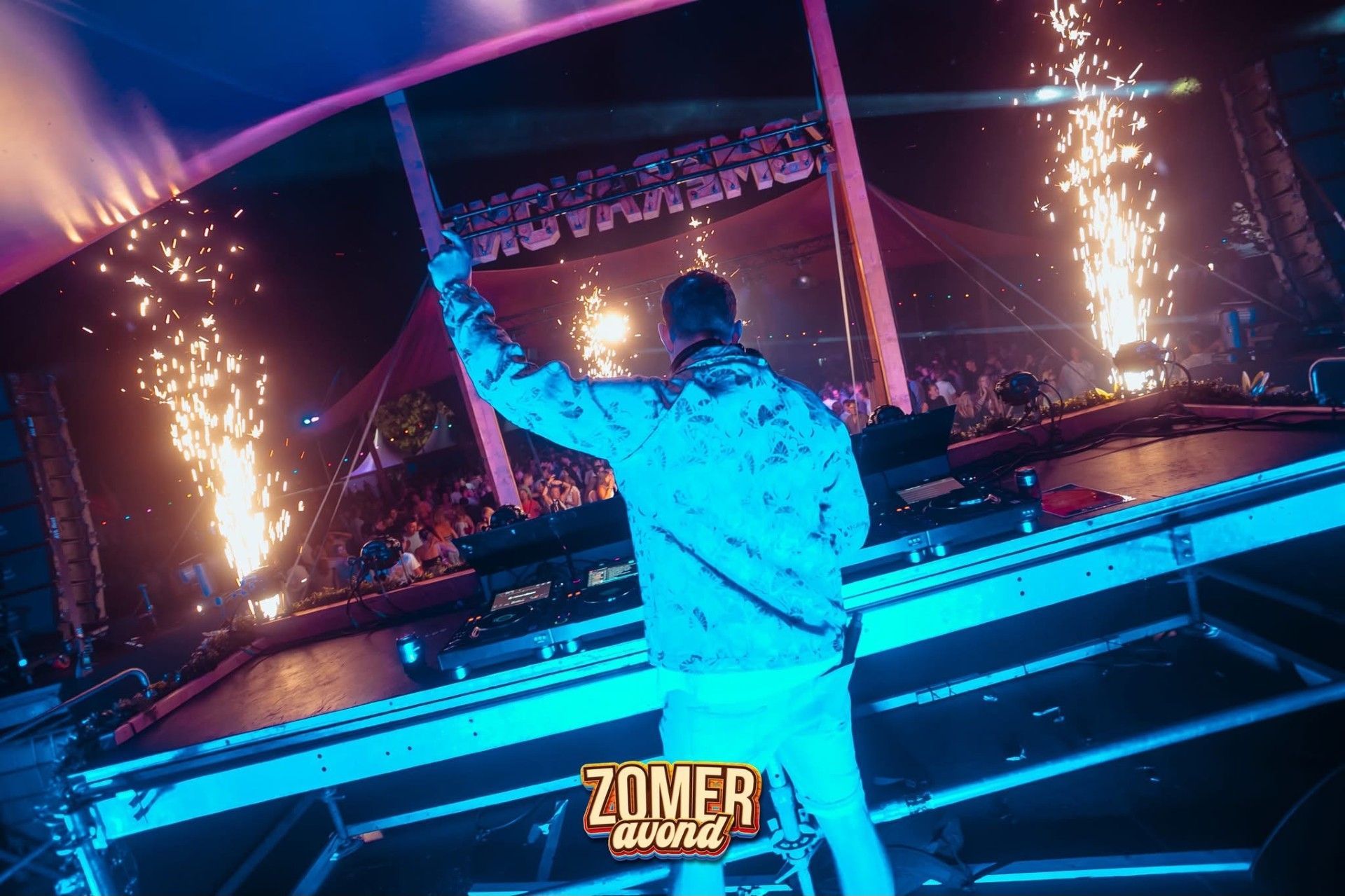 DJ treedt op op een neonverlicht podium met vonken die van beide kanten afschieten tijdens een zomerevenement.