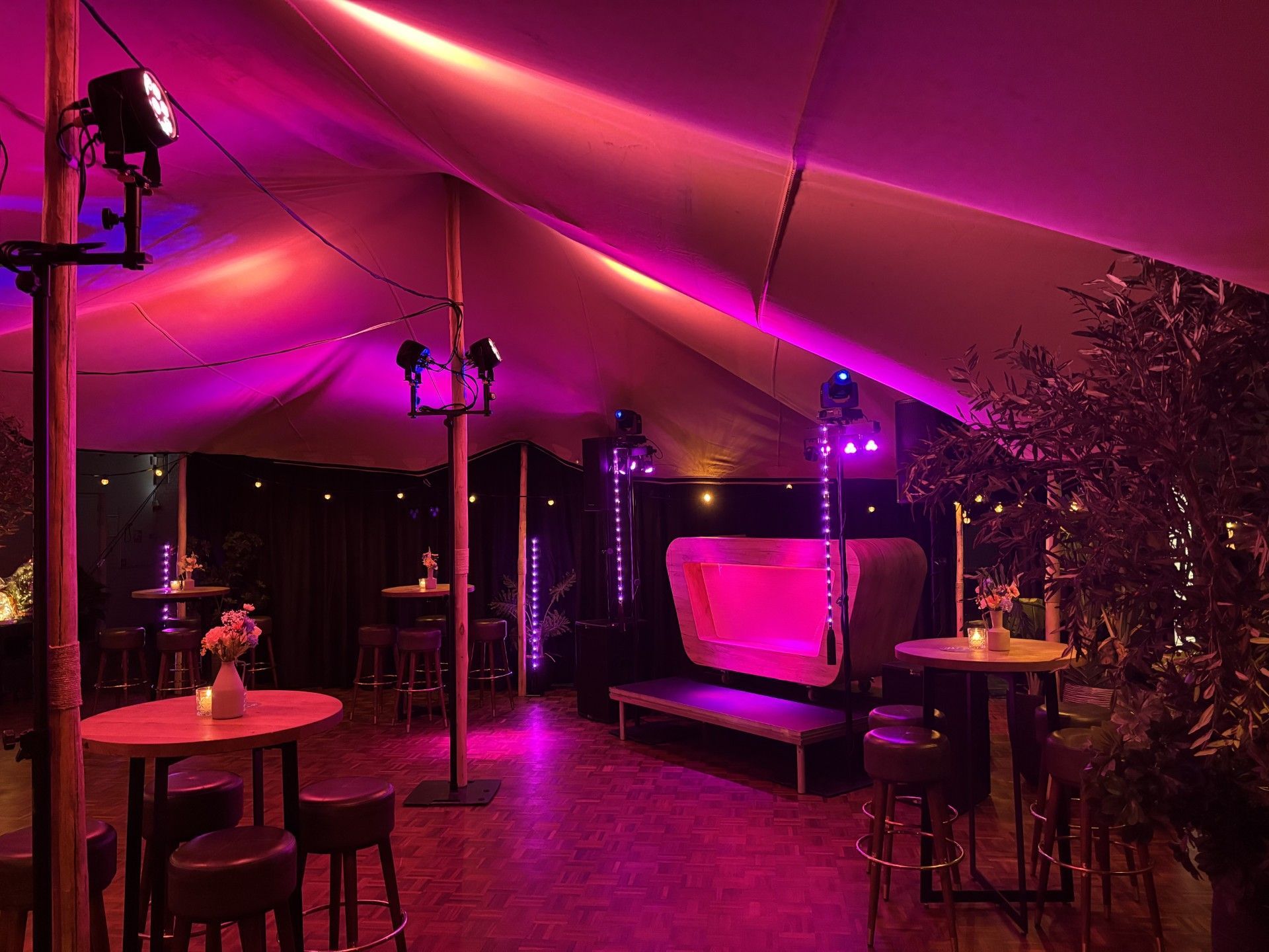 Paars verlichte loungetent voor buiten met tafels, krukjes, lichtslingers en een roze bank.