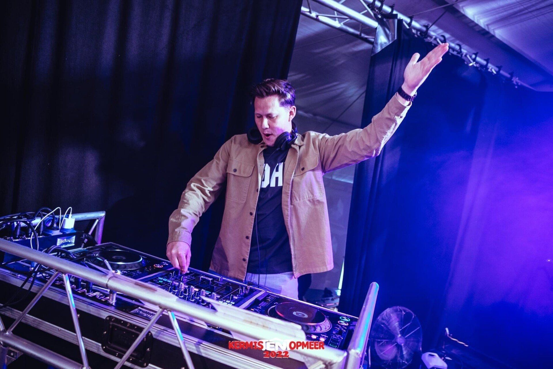 DJ steekt één hand op achter een verlichte mengtafel op een paars podium.
