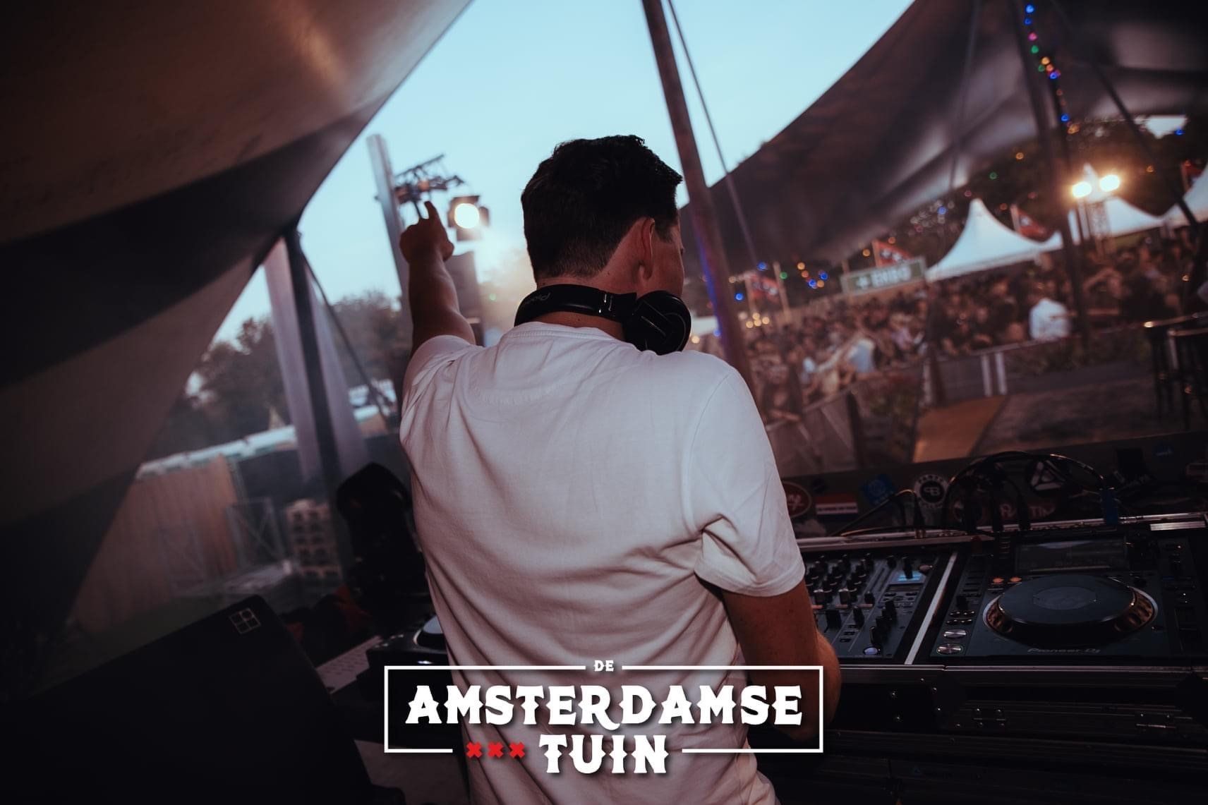 DJ die optreedt op het podium van het Amsterdamse Tuin festival, van achteren gezien met publiek en tenten op de voorgrond.
