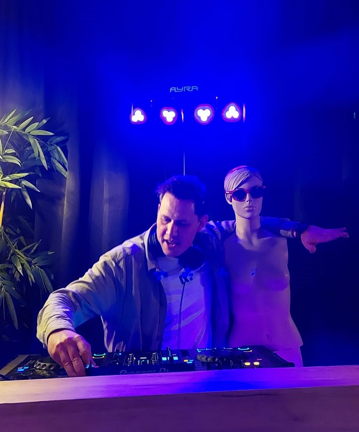Twee dj's draaien achter een mengpaneel onder blauwe podiumverlichting, de een mixend en de ander wijzend.