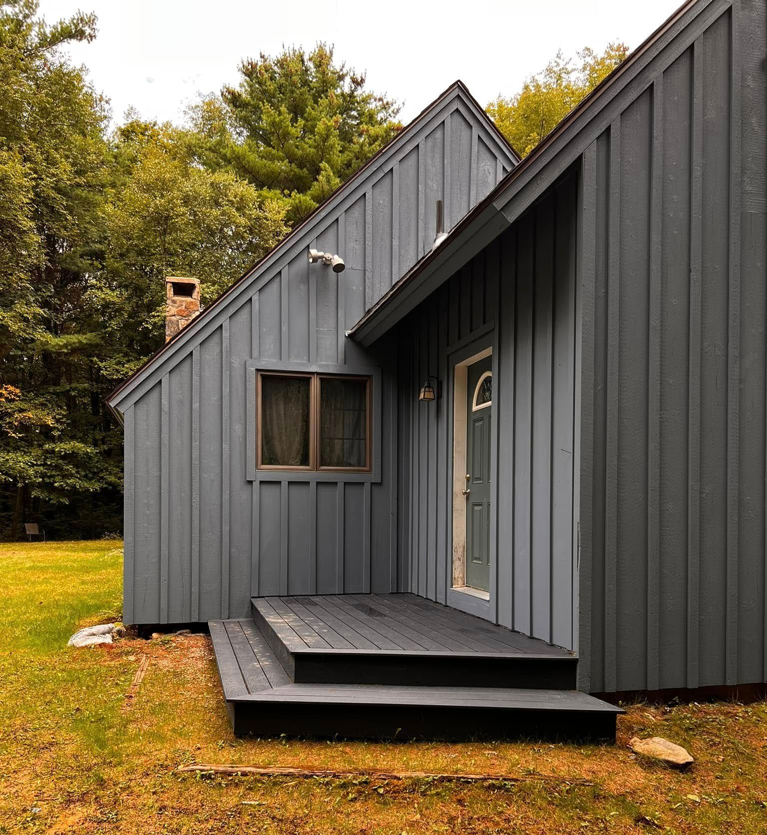 A blue, vertical-sided house with a steep roof, a small wooden deck, and a single door, set against a wooded backdrop.