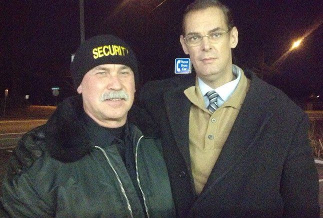 Two men standing together at night, one in a black security hat and jacket, the other in a dark coat and tie.