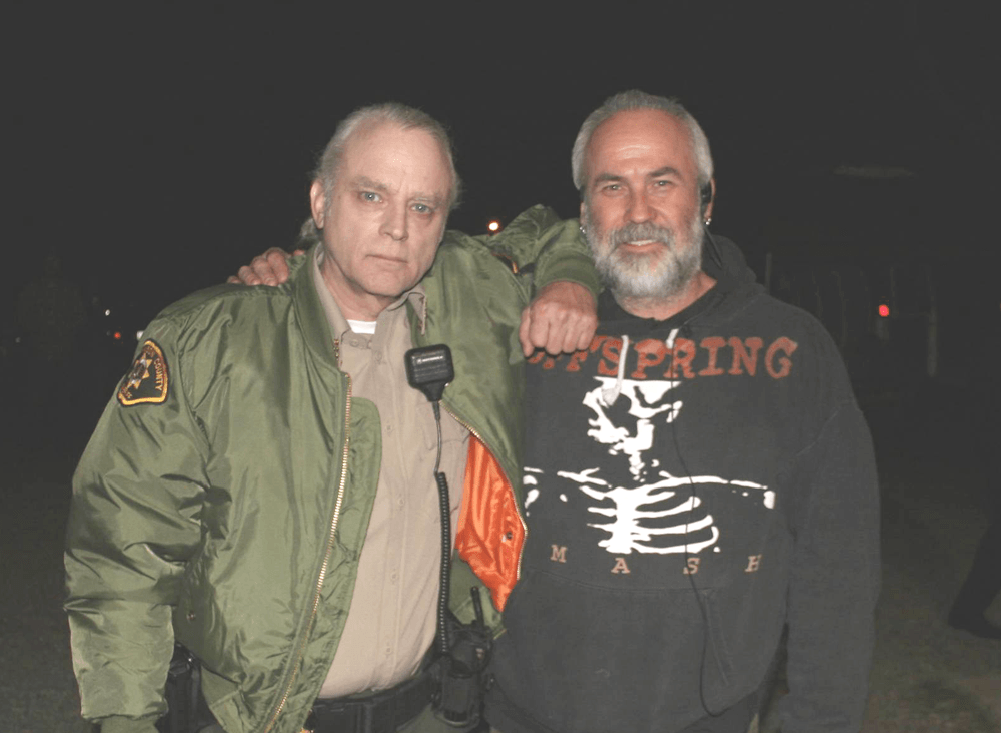 Two men posing at night, one in a green jacket and one in a black shirt with an orange skeleton graphic.