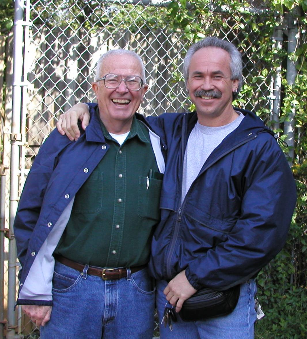 Two smiling men standing together outdoors, one with his arm around the other