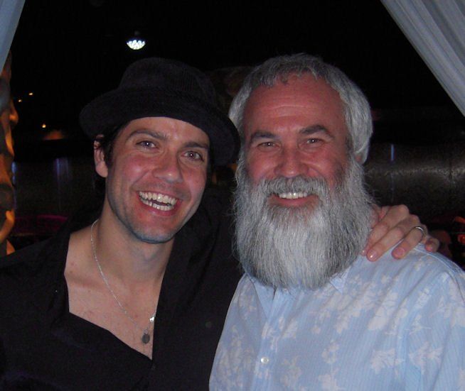 Two smiling men posing together at a dimly lit event, one in a black hat and the other with a white beard