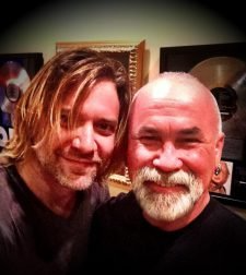 Two smiling men pose for a selfie in front of framed gold records.